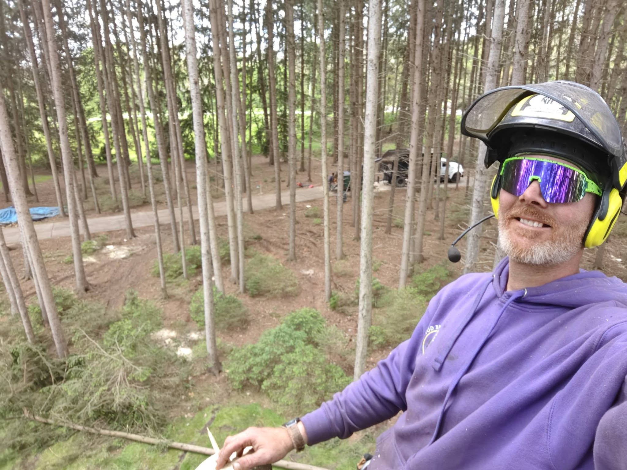 Jake selfie at the top of tall pine trees with crew and equipment visible on the ground far below